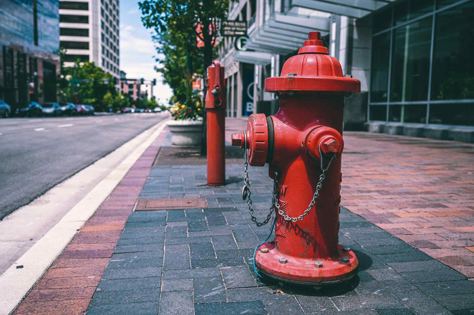 Ein roter Hydrant mit Ketten steht an einem sonnigen Tag auf einer leeren Straße in der Nähe von Straße und Gebäude.